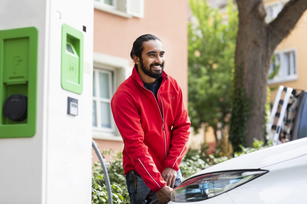 Smiling man in red jacket charging an electric car outdoors near a green station. Germany